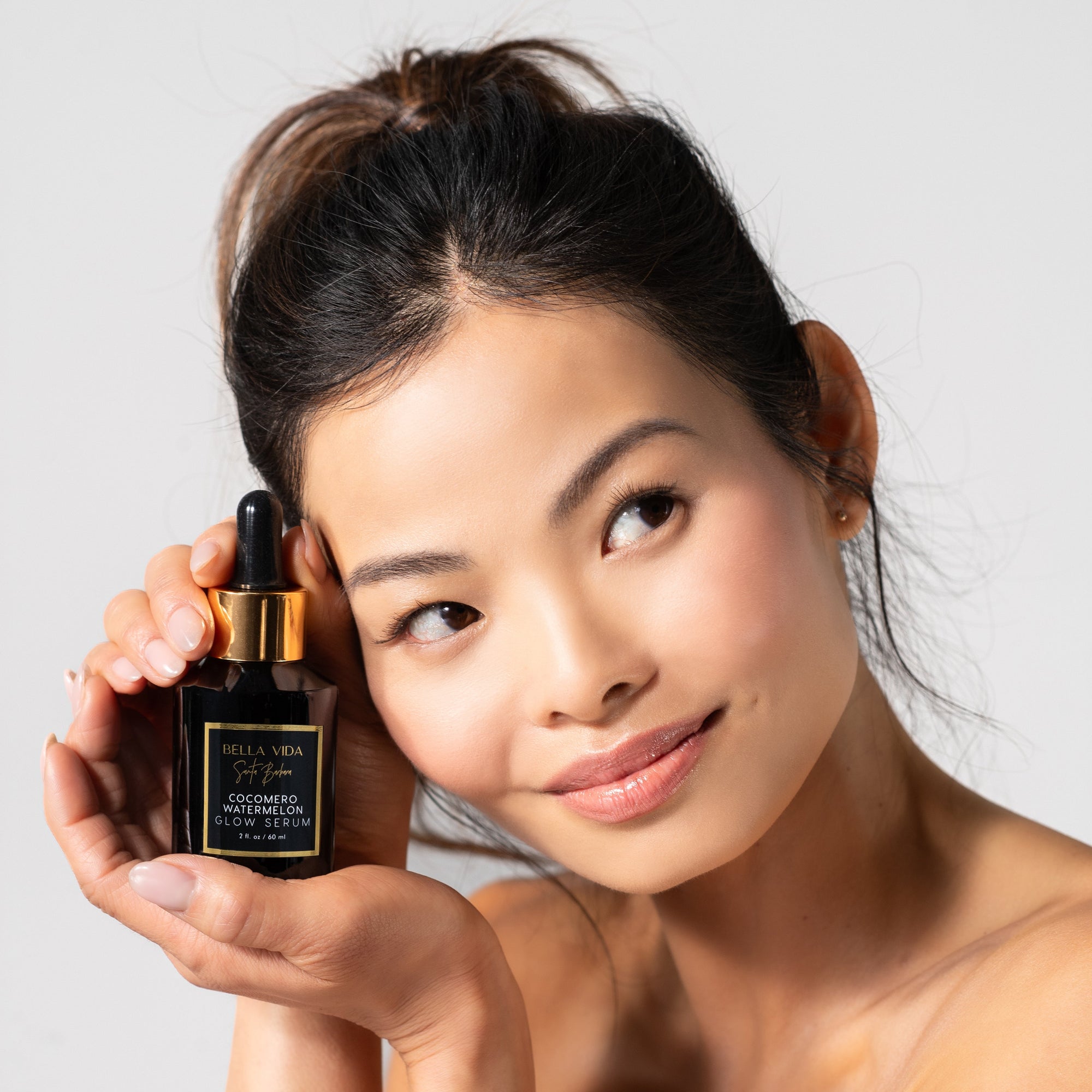 Woman holding A black bottle with a dropper of Cocomero Watermelon Glow Serum with Hyaluronic Acid, labeled with the brand Bella Vida Santa Barbara luxury skincare, and an award badge for 'Beauty Shortlist Editor's Choice 2024'. The product is described as helping to achieve a glow and radiance for the skin.