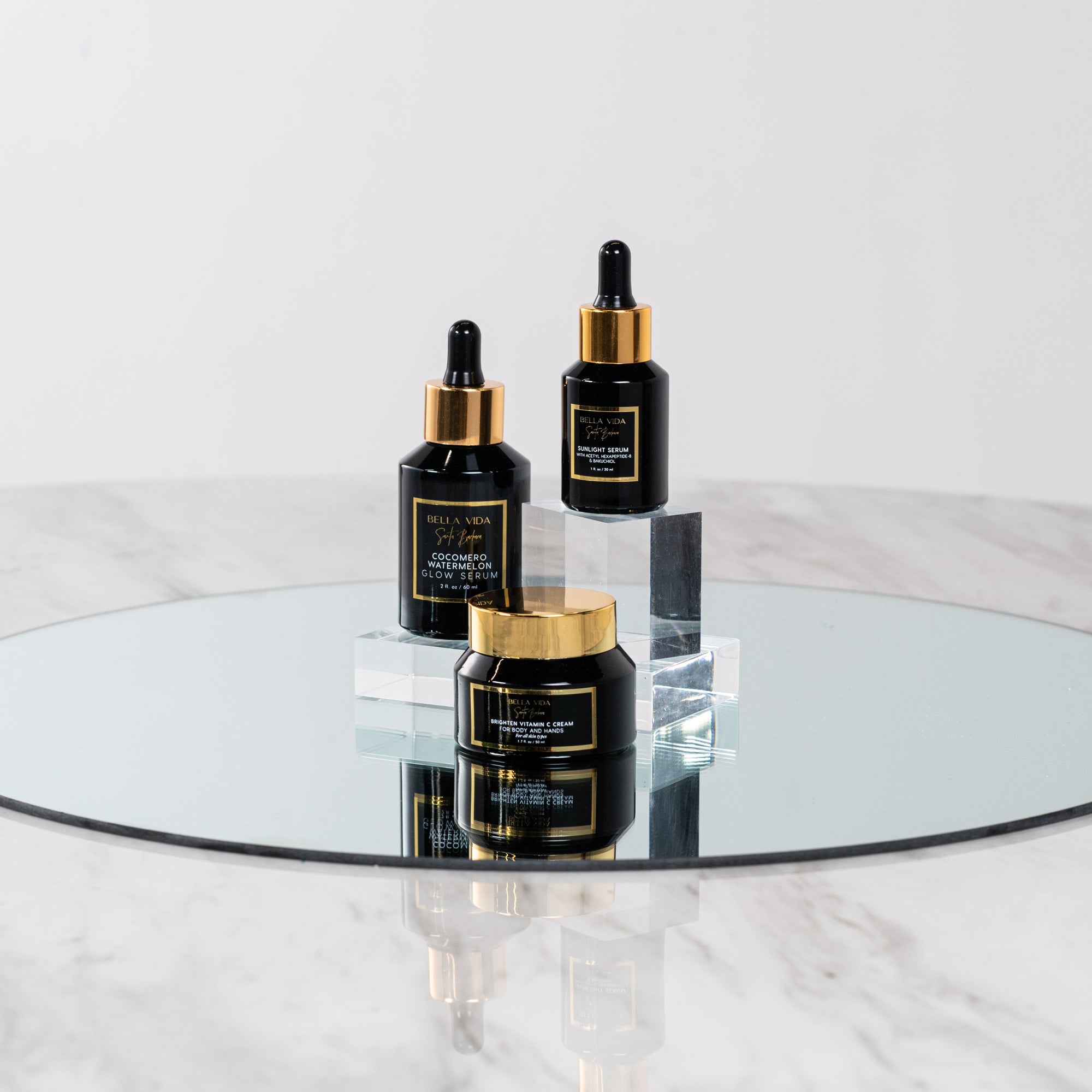 Three black Bella Vida Santa Barbara luxury skincare bottles with gold caps on a round glass table. Sunlight Serum, Cocomero Watermelon Glow Serum and Brighten Vitamin C Cream
