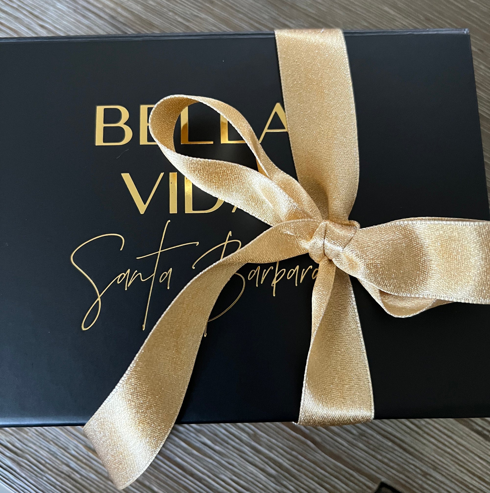 Black gift box with gold ribbon and 'Bella Vita' branding on a wooden surface